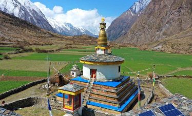 A traditional Buddhist stupa surrounded by green fields in Tsum Valley, Nepal, with snow-capped mountains in the background under a bright blue sky.