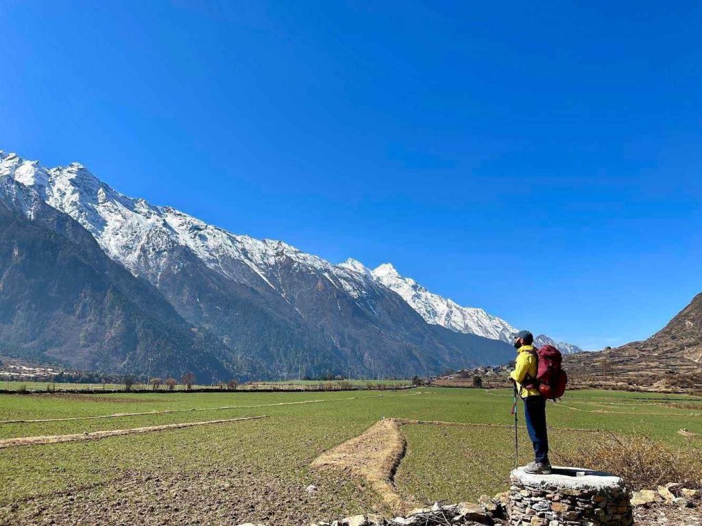 Trekker standing on a stone wall in Tsum Valley with vast green fields and snow-covered mountains.