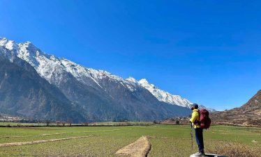 Trekker standing on a stone wall in Tsum Valley with vast green fields and snow-covered mountains.