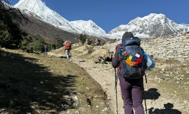 Trekker walking toward snowy Himalayan peaks on the Manaslu Circuit trail near Samagaun under clear blue sky.